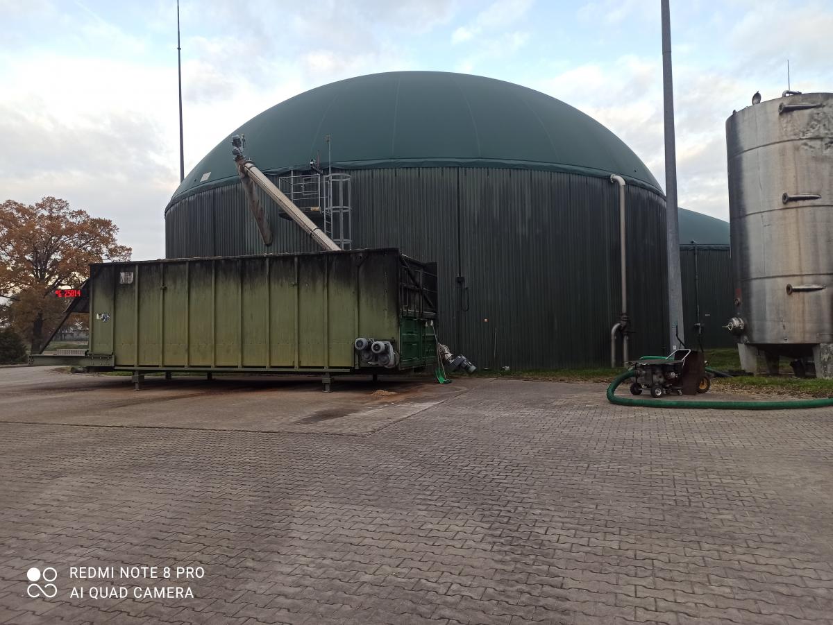 Zdjęcie przedstawia instalację biogazowni z dużym, zielonym zbiornikiem i kontenerem. Na pierwszym planie widoczna jest kostka brukowa.